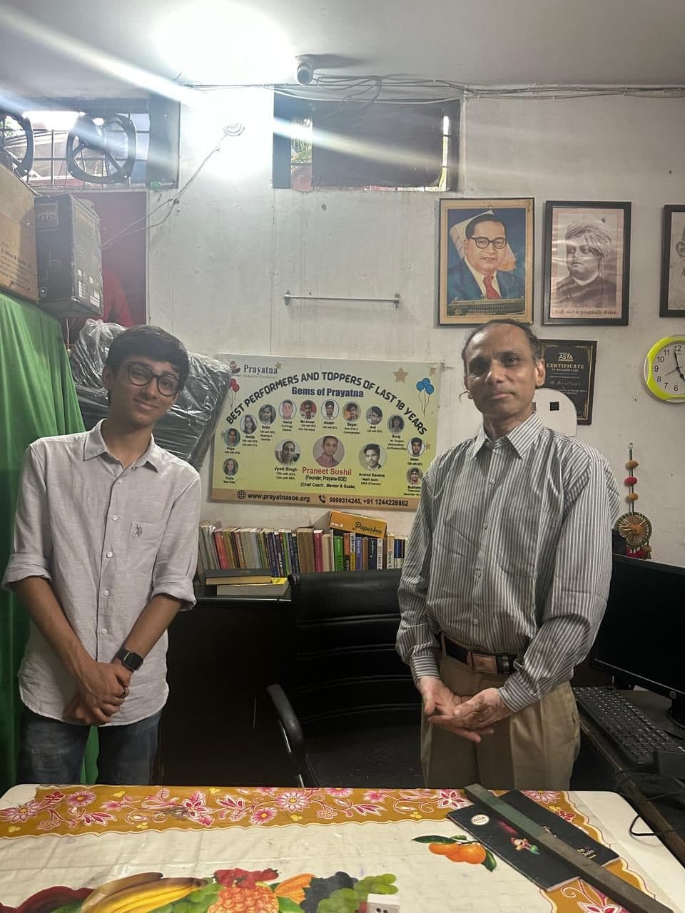 A member of the Edugram team standing with a member of Prayatna's team in the centre's office, beside a board listing the centre's top-performing students.